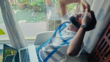 an adult man experiencing headache while working on laptop near window side at home.