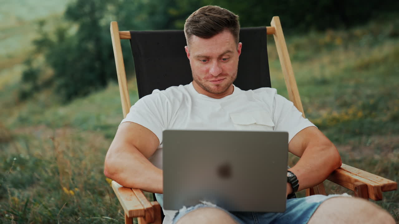 Freelancing Caucasian man works outdoors. Man sits in a folding chair focused on the laptop screen.