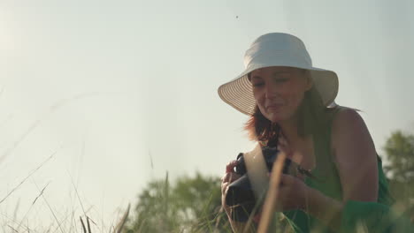 woman in green dress holding camera approaches wild plant in field during golden hour, sunlight casting soft glow as she prepares to take photograph in peaceful natural setting under clear sky