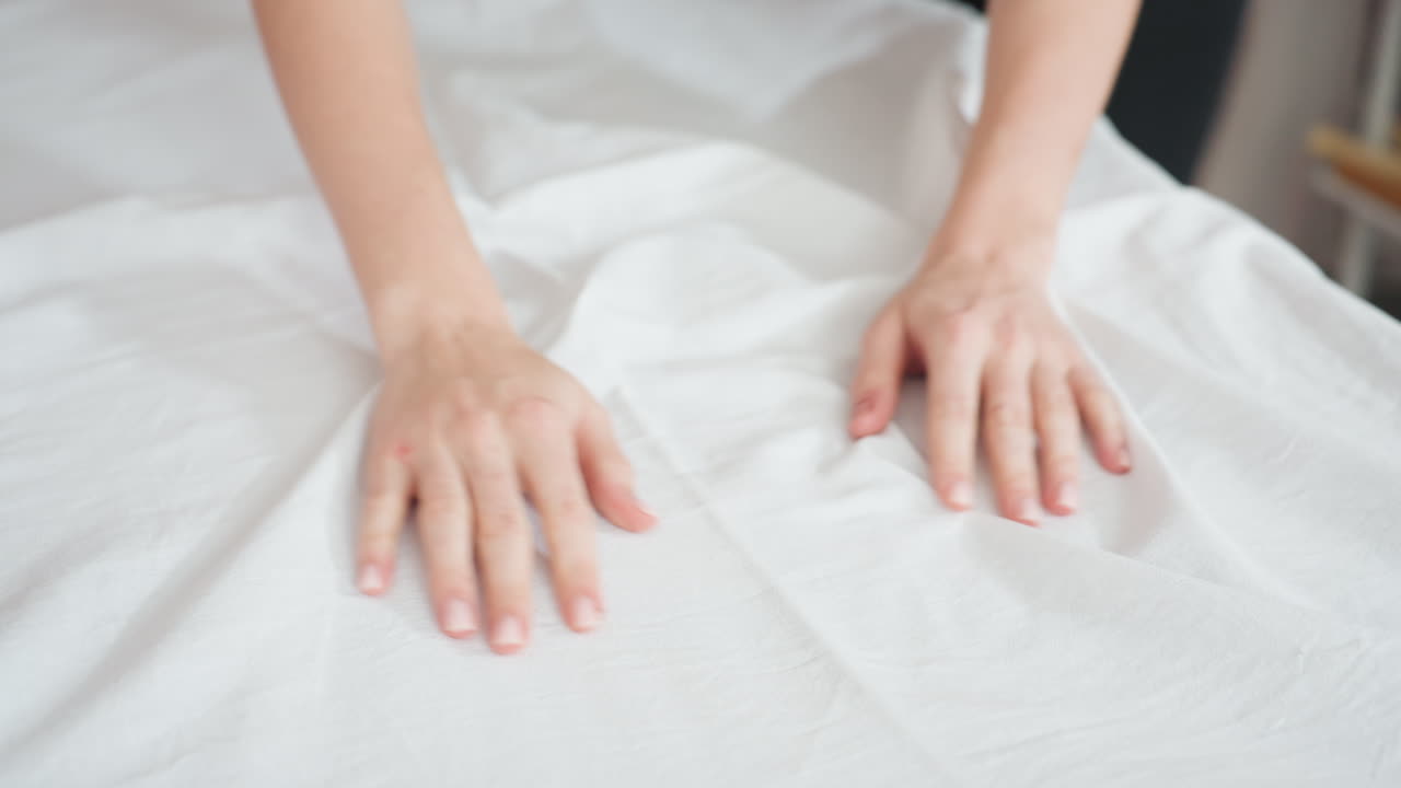 Light skin woman using both hands to smooth white bed sheet on massage table, gently removing wrinkles with focused motion in well-lit spa room, highlighting clean setup and tactile care