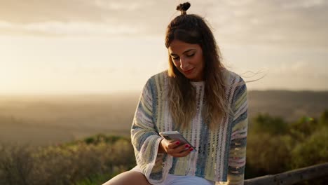 beautiful woman sitting, using smartphone outdoor with sun in the background at sunset