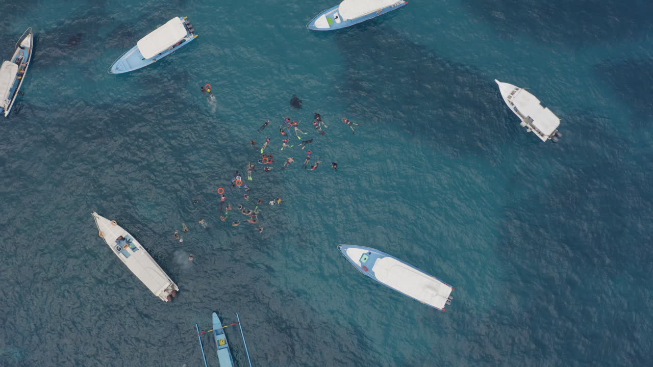 Group of tourists snorkeling in blue water at the coral shoals, surrounded by tourist boats, Nusa Penida, Bali, Indonesia. Aerial shot