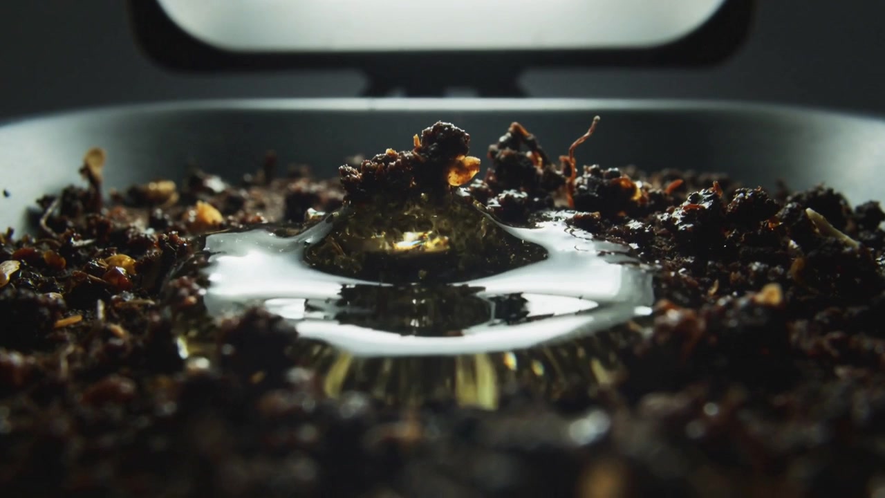 Gravity pulling amber-tinted droplet falling and spreading on shallow tray, lifting dark granules