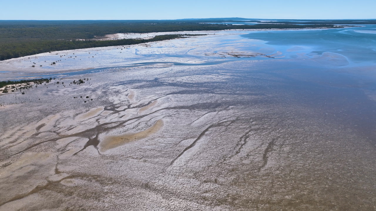 Aerial cruises over the dazzling scene of channels, mud flats, and dotted mangroves at low tide. Capturing bright iridescent reflective qualities from the intense sun, Tin Can Bay, Queensland.