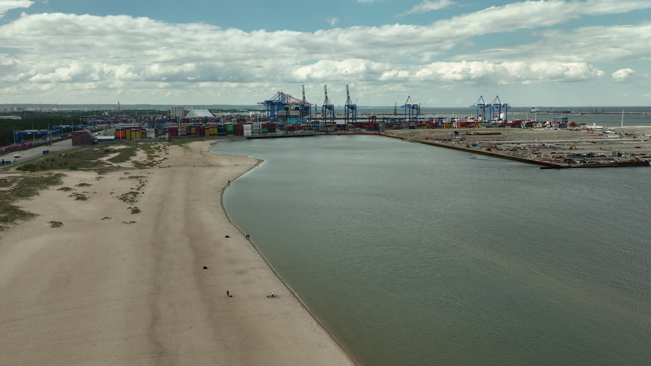 Aerial view of the Port of Gdańsk, showcasing the industrial area with cranes, containers, and a sandy beach along the shoreline under a cloudy sky