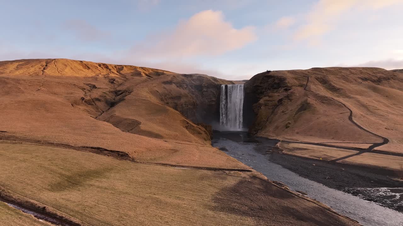 Golden hour drone pushing toward Skógafoss waterfall, Iceland, volcanic hills, winding riverbed, soft mist and warm sunset glow.