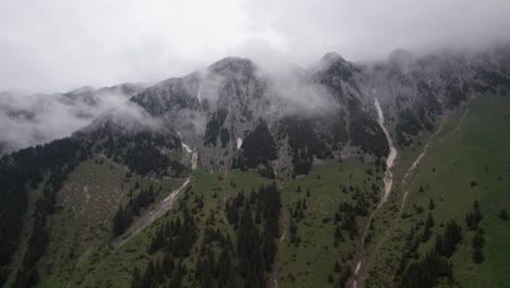 Misty-mountain-peaks-with-drifting-clouds-in-a-lush-green-valley,-aerial-view