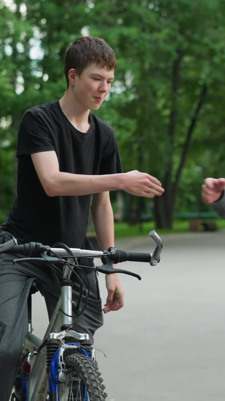 두 명의 친구가 자전거를 타고 공원 환경에서 상호 작용하며, 회색 을 입은 사람은 친구가 손가락 프로 그를 환영하기 위해 올라오는 동안 기다리고 있으며, 배경은 흐릿하고, 울창한 녹색 나무를 보여줍니다.