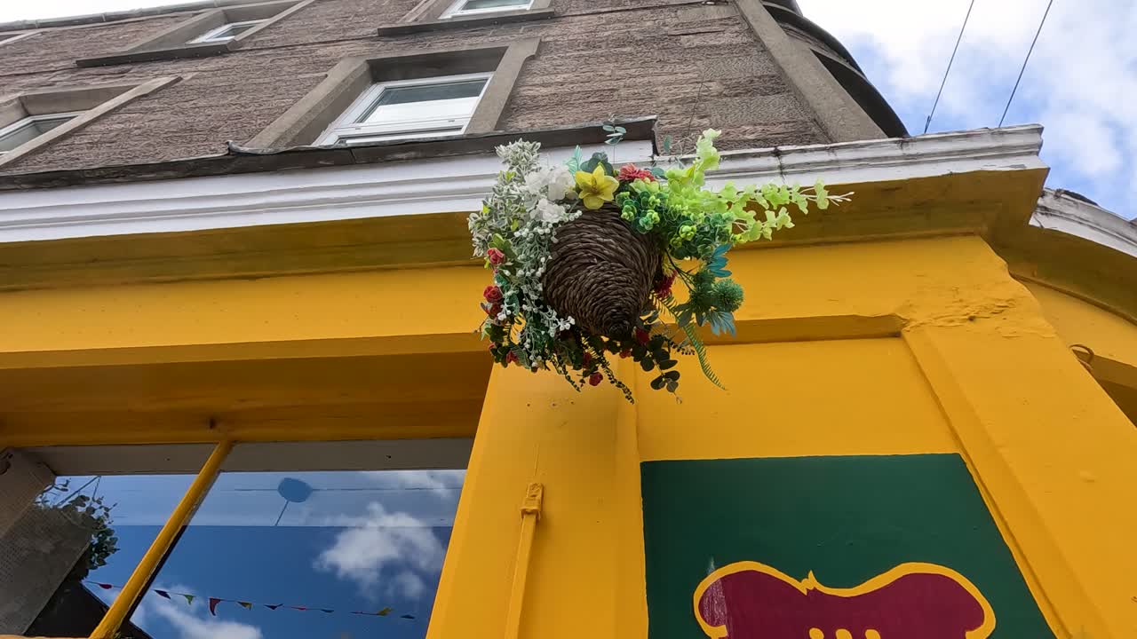 A hanging flower basket gently sways in the wind outside a bright yellow storefront in Broughty Ferry, Dundee, under natural daylight with a low upward camera angle