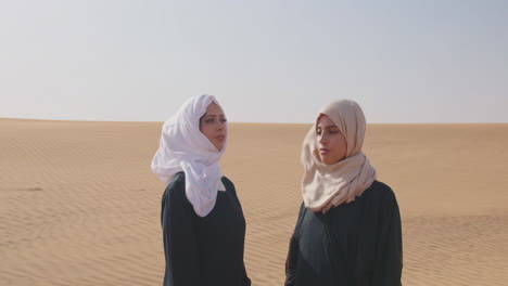 two muslim women wearing traditional dress and hijab standing in a windy desert