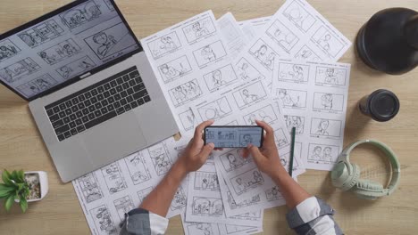 vista superior de cerca de las manos del artista masculino sosteniendo un teléfono inteligente para comprobar su concepto de dibujo en la mesa con papeles de storyboard, portátil y auriculares en el estudio