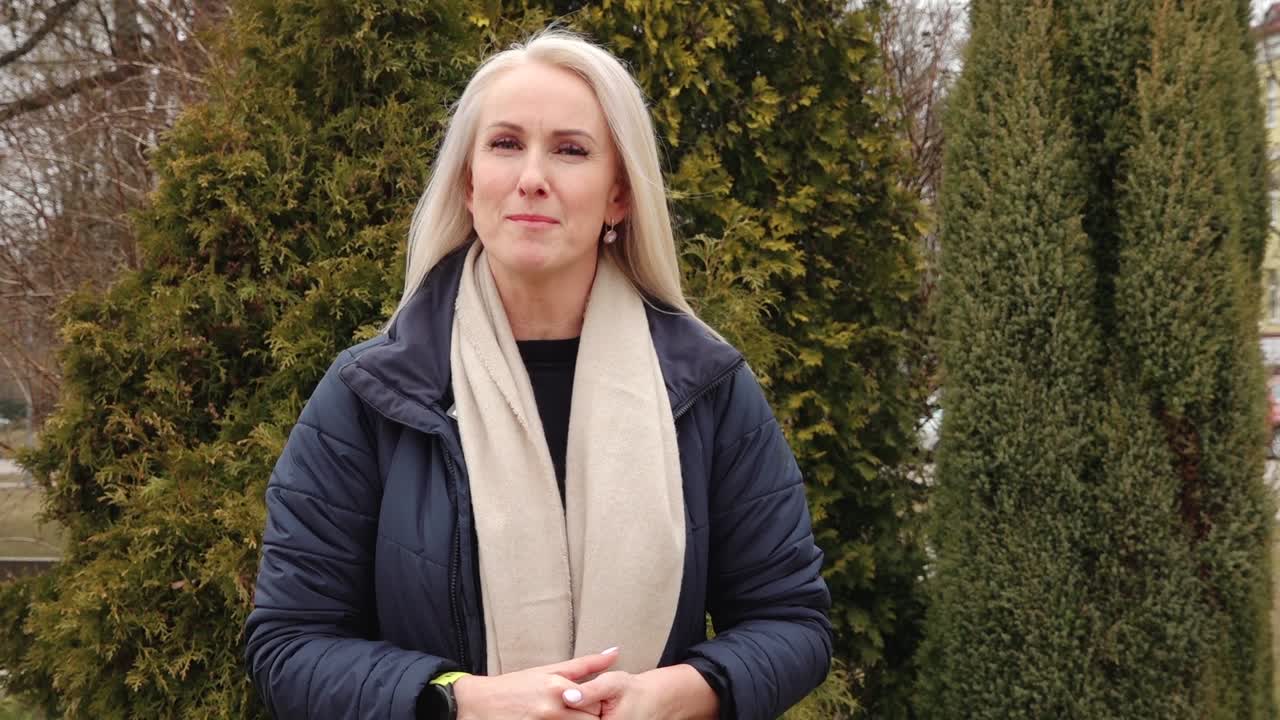 A woman with long blonde hair smiles and speaks while standing outdoors in front of evergreen trees, dressed warmly in a navy coat and beige scarf.