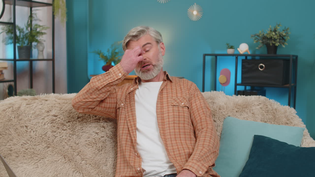 Mature old man sitting on sofa rubbing eyes with hands showing fatigue after long work at home