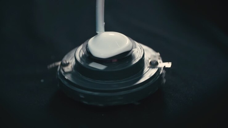 White liquid being dispensed from a nozzle, applied onto a circular speaker component, Industrial part of the assembly process for a speaker driver