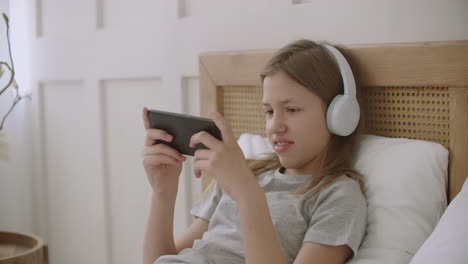 school girl is playing video game in smartphone by internet using headphones and lying on bed at vacation time