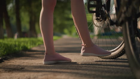 leg view of individual checking air pressure of bicycle tire with foot, featuring pink sneakers, background showcases lush greenery and trees, along with a distant building