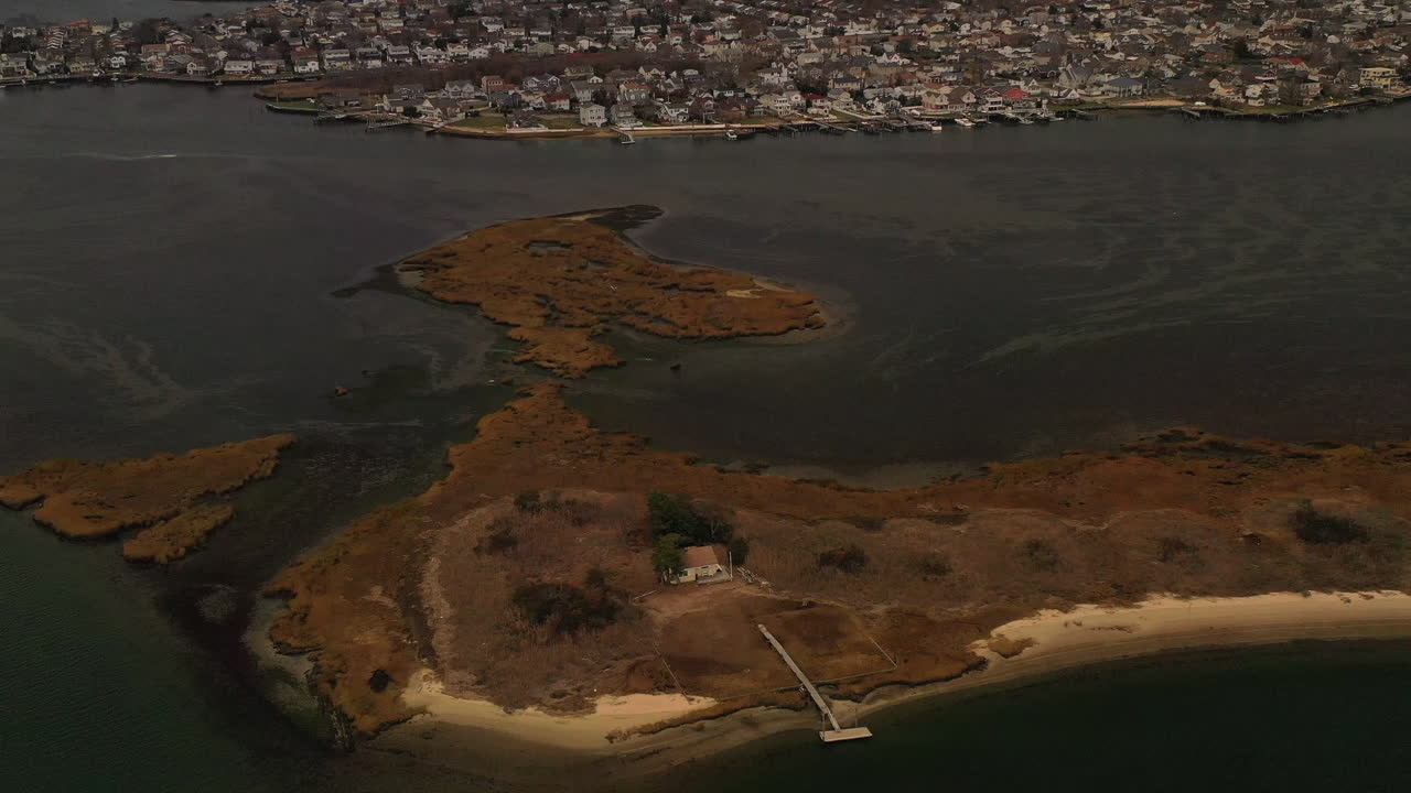 A high angle aerial view over an island with a single house - a narrow wooden pier. The camera pans right, tilting down towards the island in the salt marsh. It was taken on a cloudy day.