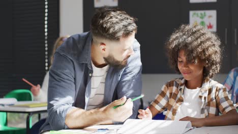 Vielfältiger-Männlicher-Lehrer-Und-Glückliche-Schulkinder,-Die-Im-Klassenzimmer-Am-Schreibtisch-Sitzen