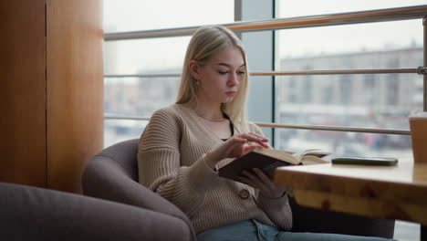 dama contemplativa da la vuelta a una nueva página mientras lee un libro pensativamente en un café con vista borrosa de la ciudad visible a través de un panel de vidrio, taza de café y teléfono descansando en la mesa