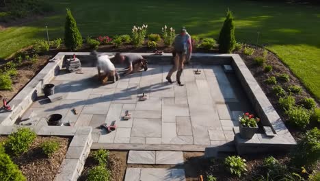 workers are installing a beautiful stone patio in a backyard garden. the warm summer afternoon provides a perfect setting for this landscaping project, featuring vibrant flowers and greenery.
