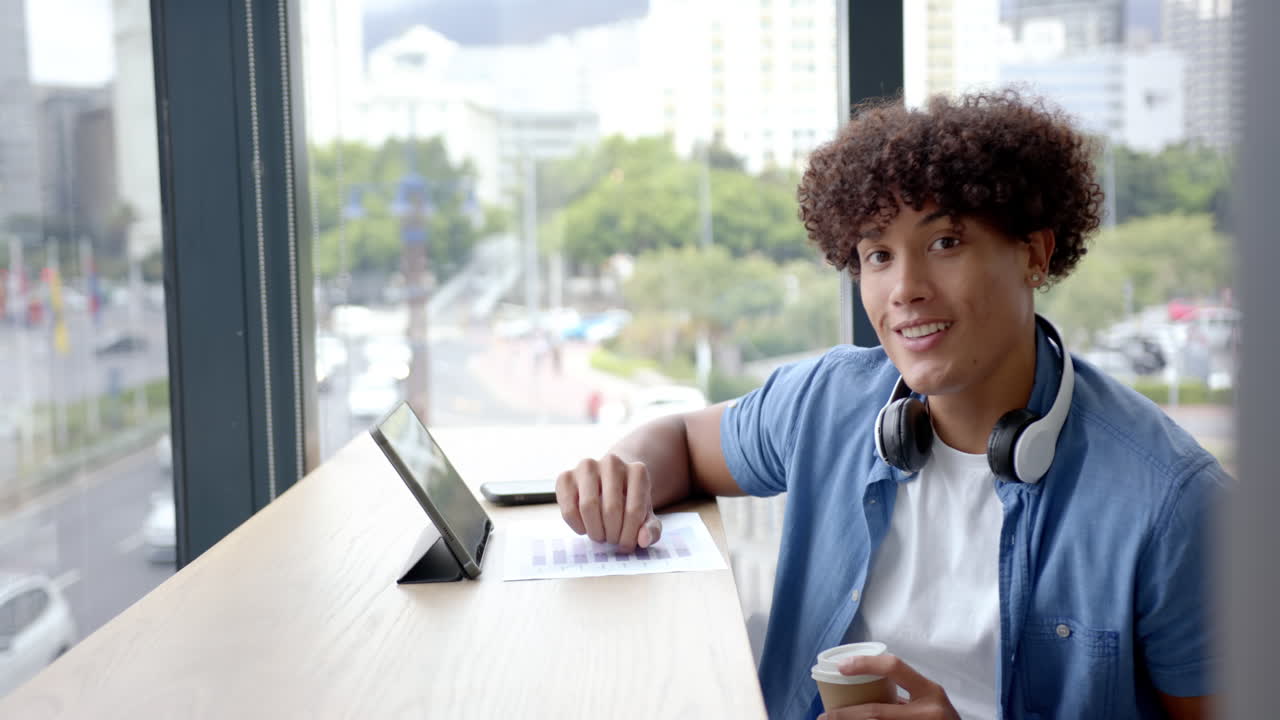 Smiling man with headphones using tablet and holding coffee in modern office