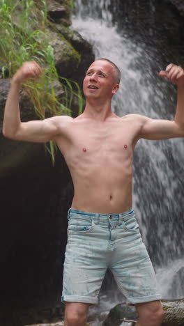 positive bare chest man enjoys vibrations of wild nature power standing near waterfall and steep cliffs at summer highland slow motion