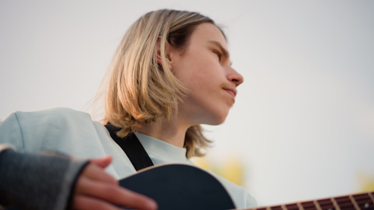 young person playing black acoustic guitar outdoors, unspecified musician gazing into distance, soft sunset light, casual sweater, finger on strings, intimate portrait with cinematic bokeh and relaxed