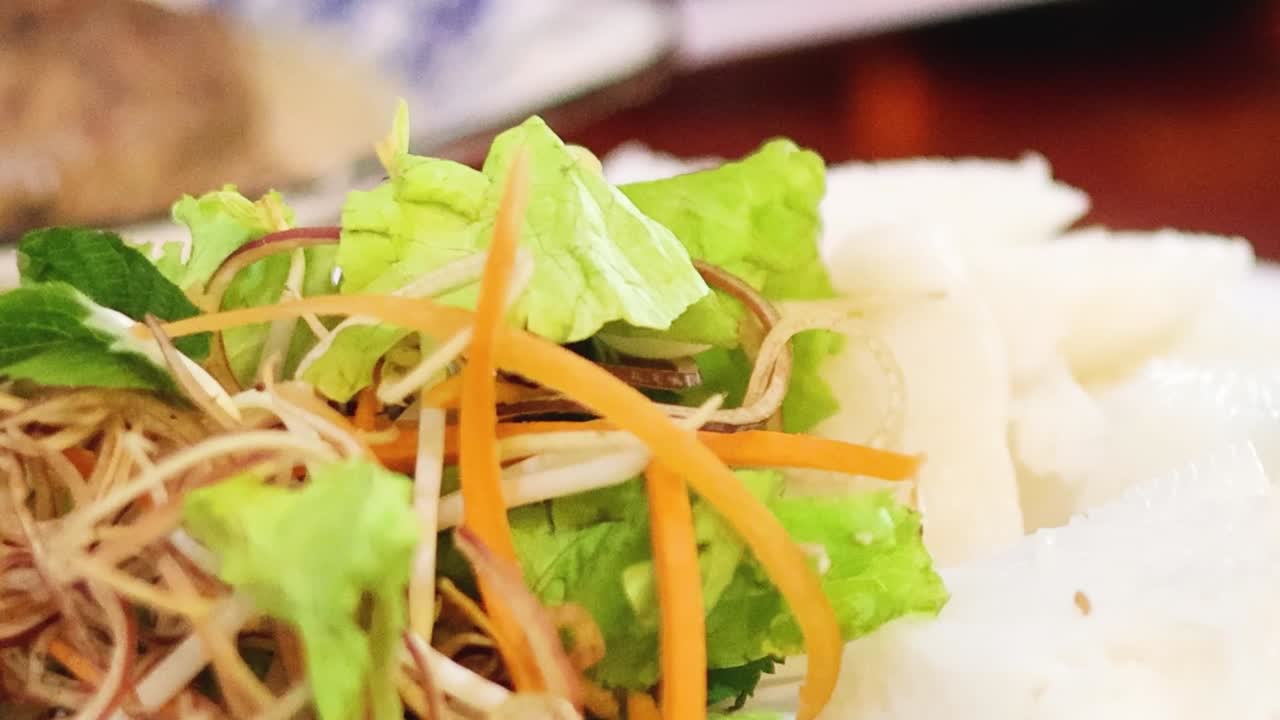 Close-up of chopsticks placing fresh herbs and vegetables on rice noodles.