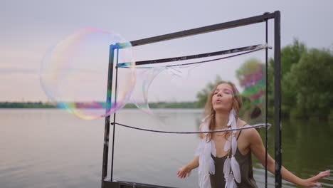 una joven artista sopla muchas burbujas de jabón muestra un espectáculo teatral usando un marco en cámara lenta al atardecer en un lago