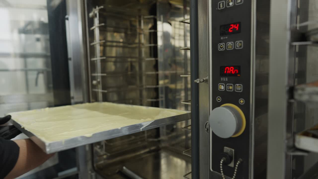 мужчина вручную кладет поднос с сырым тестом в коммерческую духовку и закрывает кулинарный инструмент. непризнаваемый профессиональный кавказский пекарь, работающий на кухне, выпекающий кондитерские изделия. профессия и кухня.