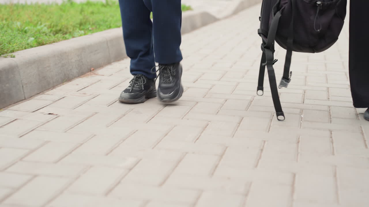 Para siswa berjalan di trotoar dengan ransel bersiap untuk perjalanan ke sekolah, tampilan dekat kaki dan sepatu melangkah di sepanjang trotoar dekat tepi jalan, latar kampus perkotaan, seragam kasual dan tali tas terlihat