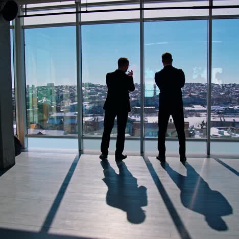 businessmen in dark suits talking during break. office workers standing near large windows and looking on city. rear view