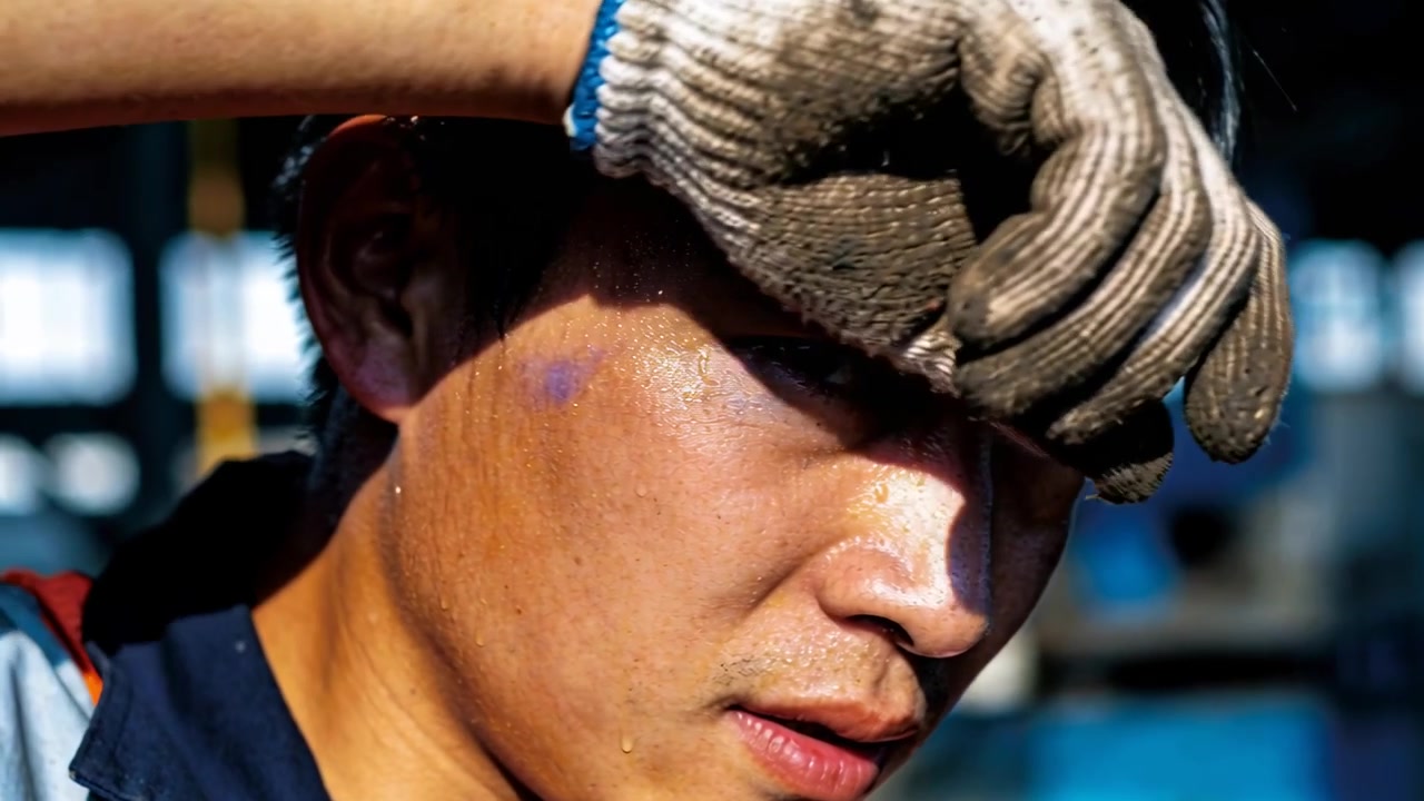 Shielding Asian worker from sun at dock adjusting grey glove, wearing navy shirt, red vest strap