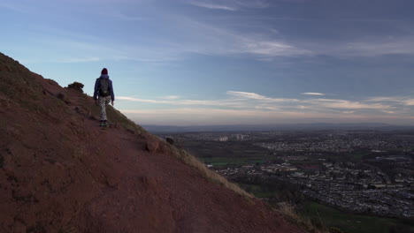 Dolly-Kamerafahrt-Eines-Mädchens,-Das-Abends-Auf-Dem-Wanderweg-Den-Arthurs-Seat-Mountain-Hinaufsteigt,-Dämmerung-Mit-Der-Stadt-Edinburgh-Im-Hintergrund