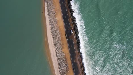 Aerial drone shot from above, capturing a road bisected by ocean waters on both sides, with waves splashing onto the surface.