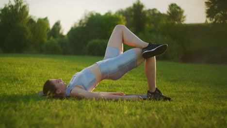 Mujer-Joven-Haciendo-Levantamientos-De-Cadera-Con-Una-Sola-Pierna.-Ajuste-Los-Músculos-De-Los-Glúteos-Del-Entrenamiento-Del-Atleta-Con-El-Levantamiento-De-Glúteos-Del-Puente-Del-Piso-De-Una-Pierna-En-El-Gimnasio-Al-Aire-Libre-De-Verano-En-El-Césped