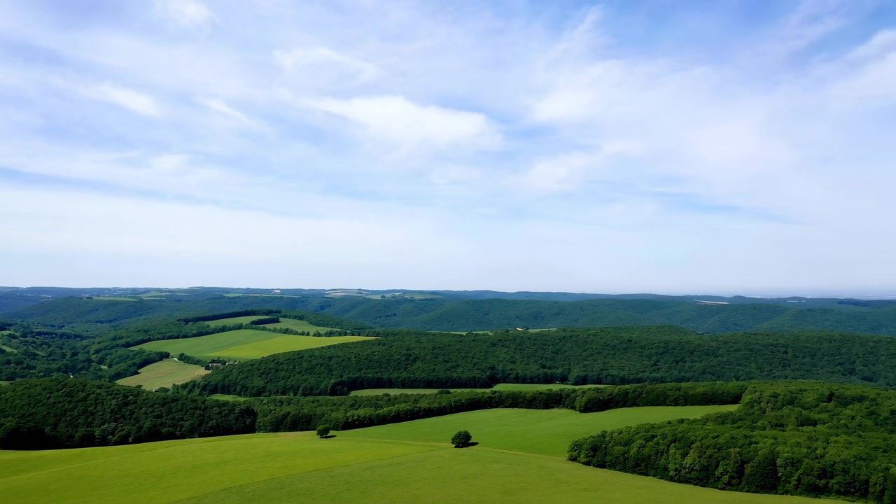 Expansive countryside reveals vivid farmland, wooded ridges, and scattered trees while smooth lateral movement follows rolling terrain under clear daylight and wispy cloud cover.