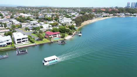 Beautiful-Gold-Coast-sunny-day,-aerial-view-of-ferry-on-canal-passing-luxury-million-dollar-homes,-Queensland-Australia