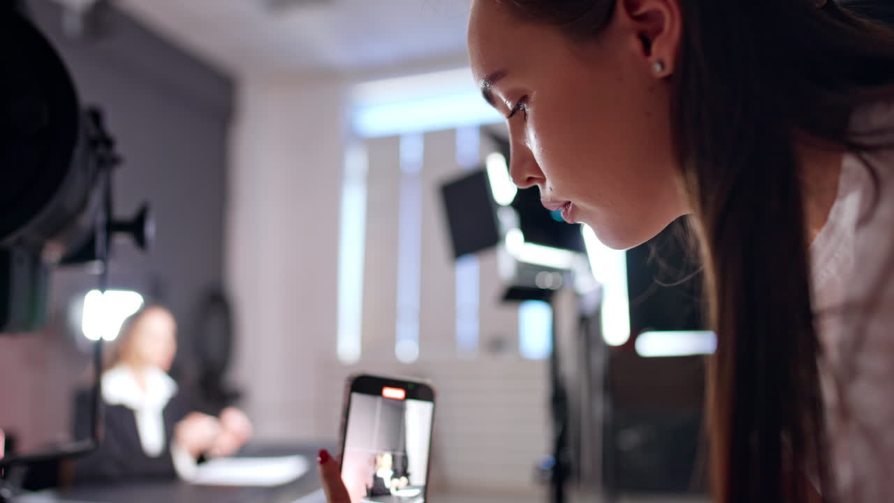 Caucasian brunette holds a smartphone taking video. Woman at blurred backdrop talks to camera. Studio footage.