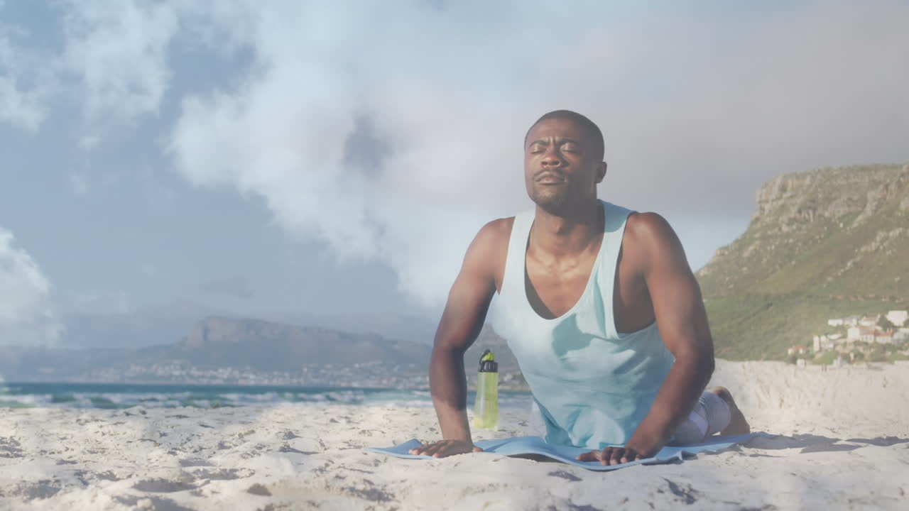 Animation of clouds over african american man doing cobra yoga pose on beach
