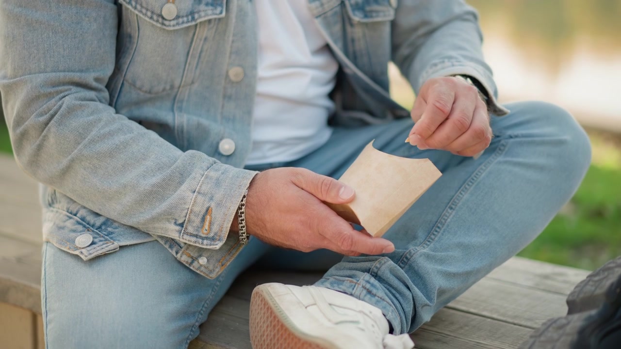Primo piano di un uomo in giacca di jeans e jeans che apre un pacchetto di carta marrone rivelando un alimento. L'uomo tiene uno snack avvolto e indossa un braccialetto a catena d'argento al polso. Ambientazione esterna con vegetazione sfocata.