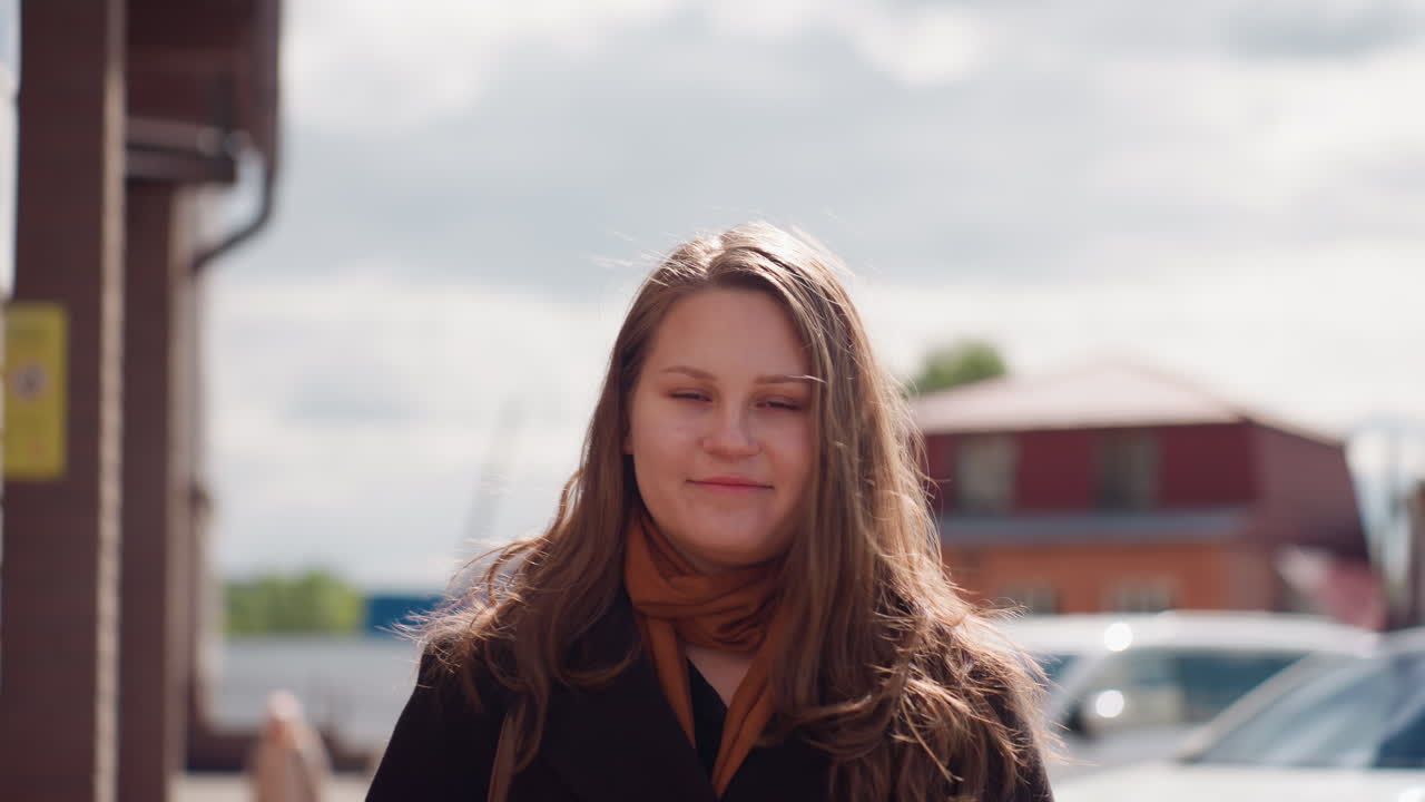 Joven con abrigo se levanta las gafas de sol con una cálida sonrisa, la brisa mueve su pelo, la luz del sol es suave en su rostro, rascacielos de la ciudad y coches aparcados detrás, ambiente callejero capturado en un momento espontáneo durante una tarde brillante.