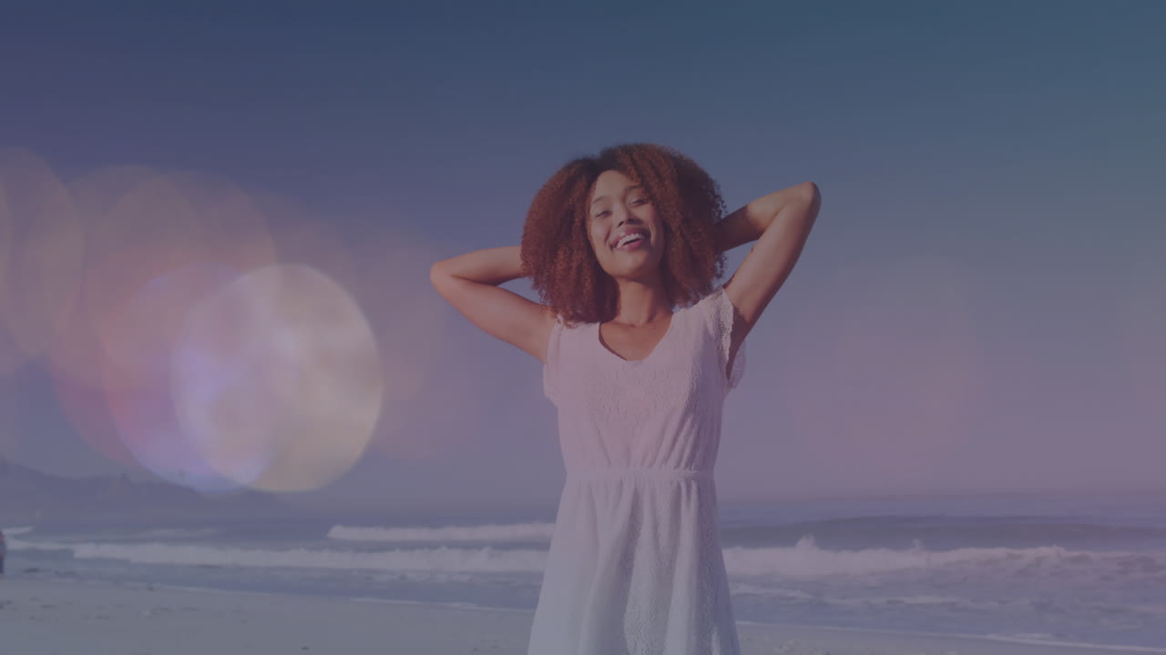 Animation of light spots moving over happy biracial woman with arms wide on beach