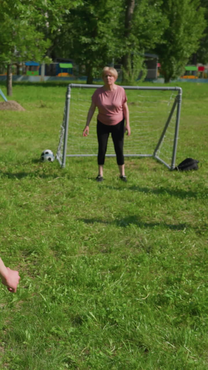 семья занимается футбольной игрой на открытом воздухе, мальчик в белом, когда он дриблирует мяч к воротам, его братья пытаются остановить его, он стреляет в мяч, мать быстро выбивает мяч