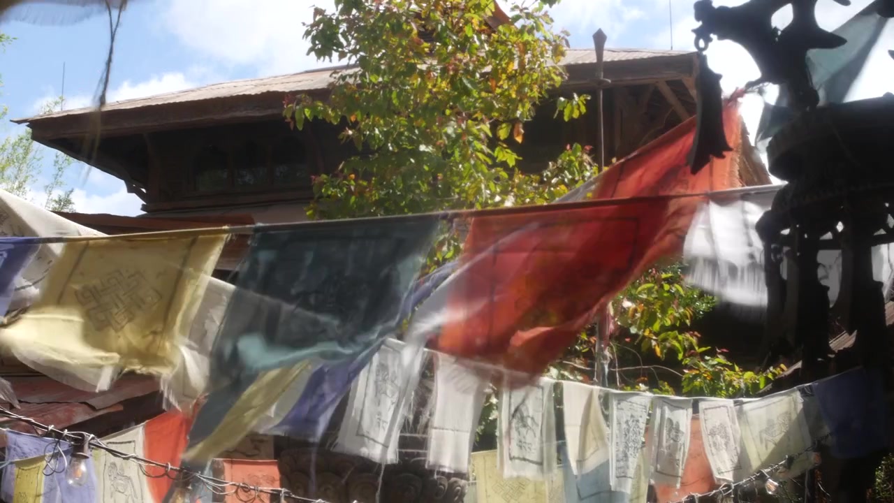 Bendera doa berkibar di samping lonceng gantung gelap di halaman kuil yang diterangi matahari. Arsitektur kayu, pepohonan rindang, dan efek buram gerakan lembut menciptakan suasana perjalanan budaya.