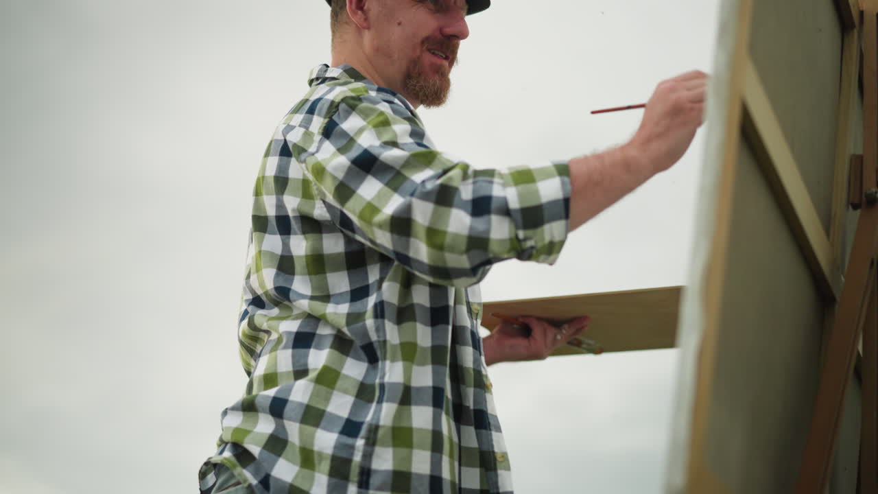A painter, wearing a black hat and checkered shirt, is captured smiling as he is fully engrossed in his painting. His joy and focus on his creative work are evident in this artistic outdoor scene