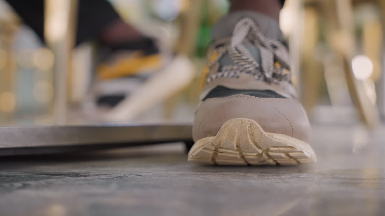 Primo piano di una sneaker sul pavimento del caffè, arrivo nervoso prima di un appuntamento, dettaglio della suola in camoscio con bokeh caldo, riflesso di piastrelle lucide e sagome di sedie, pausa intima e sottile tensione tra la coppia