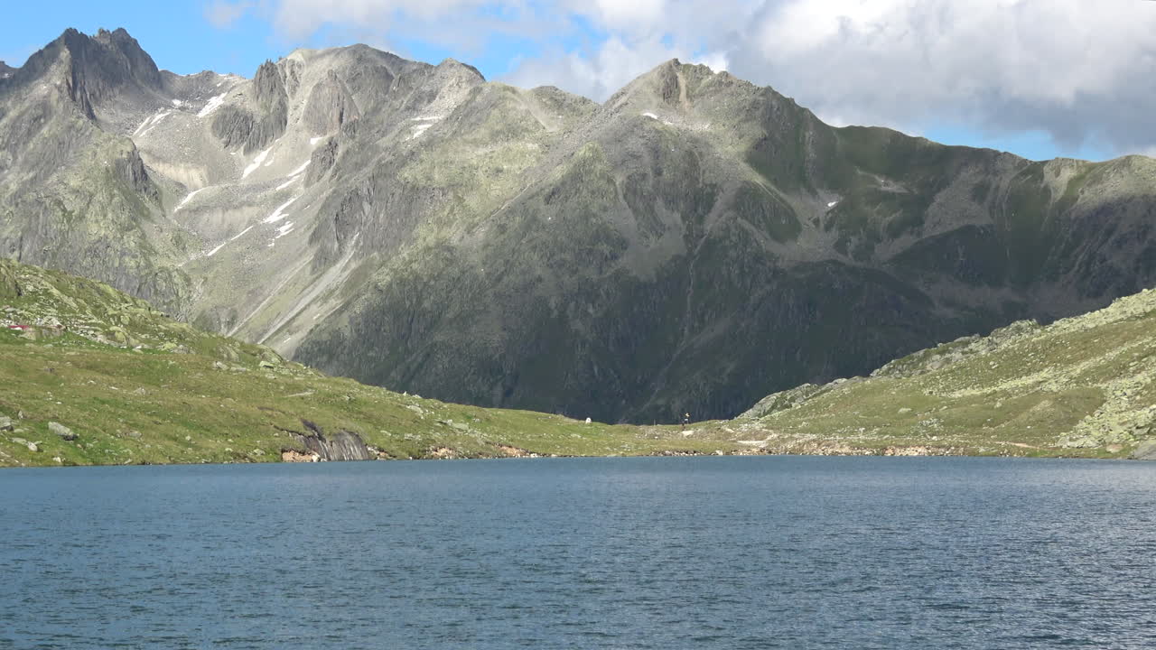 Ampia inquadratura del Lago di Merjelen in un bacino alpino con acqua blu calma, pendii montuosi rocciosi e chiazze di neve sparse sotto un cielo parzialmente nuvoloso, Svizzera.