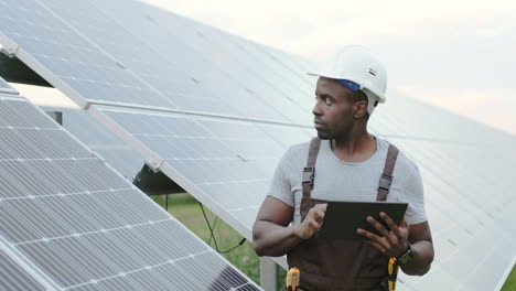 joven ingeniero afroamericano parado afuera cerca de paneles solares y usando una tableta