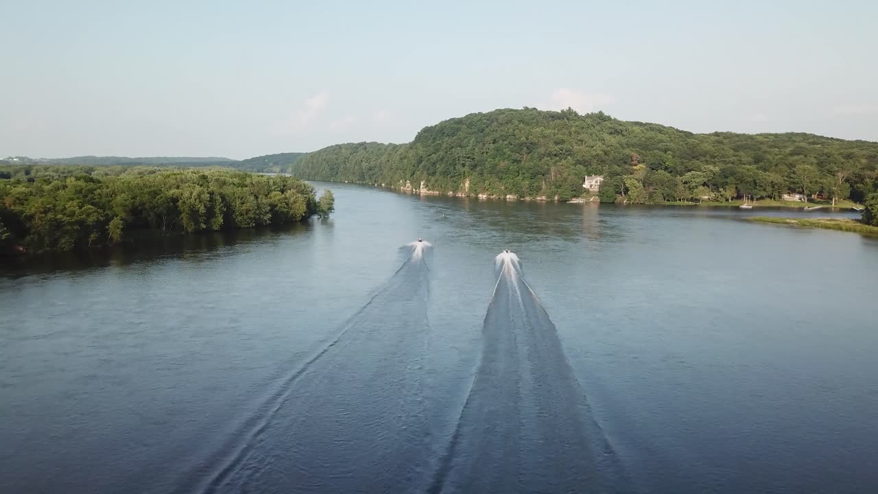Two speedboats dash side by side across a broad, tranquil river flanked by lush greenery and distant hills, their wakes trailing in perfect symmetry under clear skies.